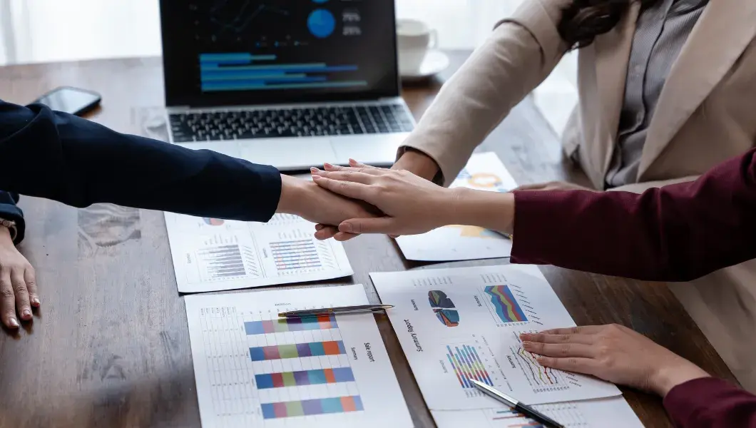 Team members stack hands over financial reports and charts on a desk, aligning on strategy